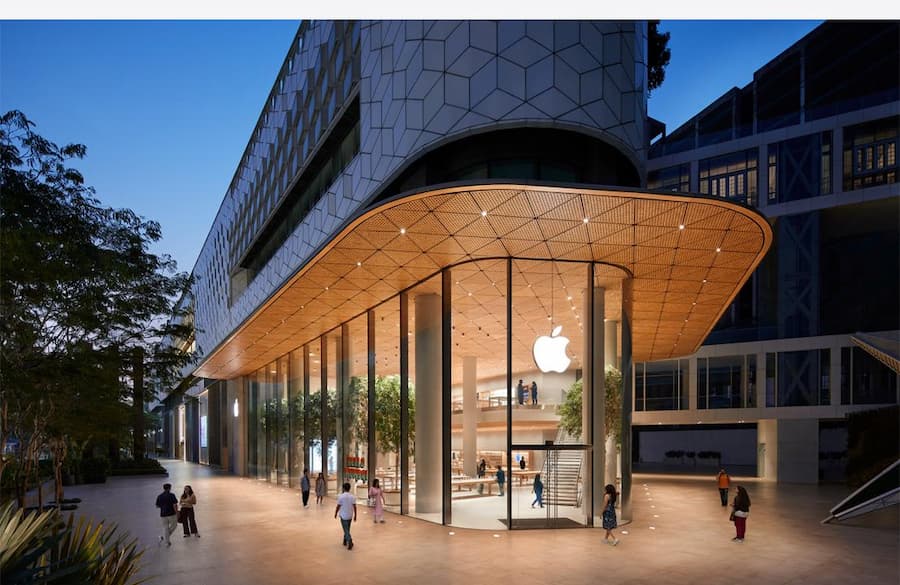 Modern Apple Store exterior at night featuring transparent glass façade, curved timber canopy ceiling, illuminated minimalist interior, and contemporary urban retail architecture