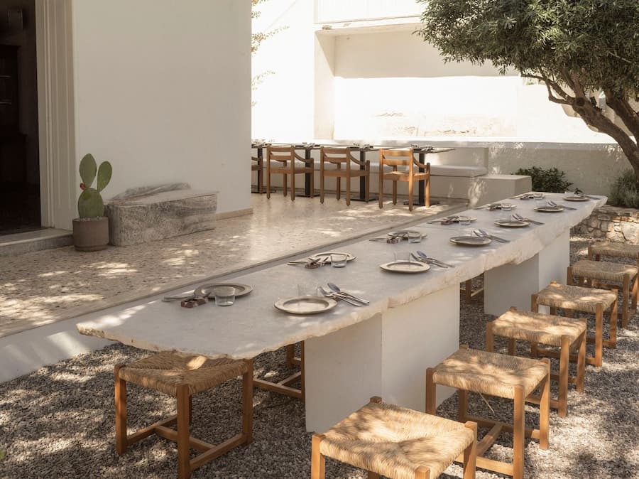 Long communal marble dining table surrounded by woven wooden stools in the Mediterranean courtyard at YAPA Antiparos