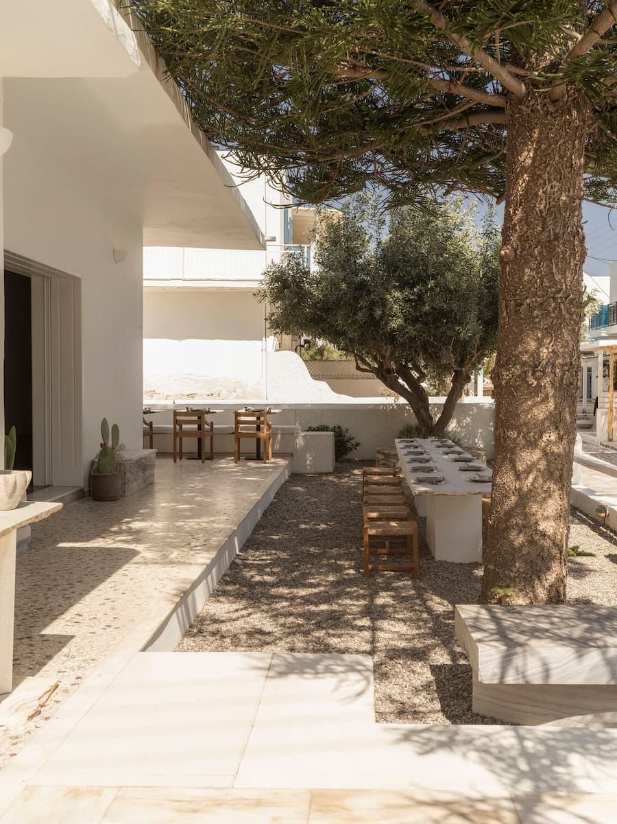 Mediterranean courtyard dining beneath mature trees with communal marble tables at YAPA Antiparos restaurant, Greece