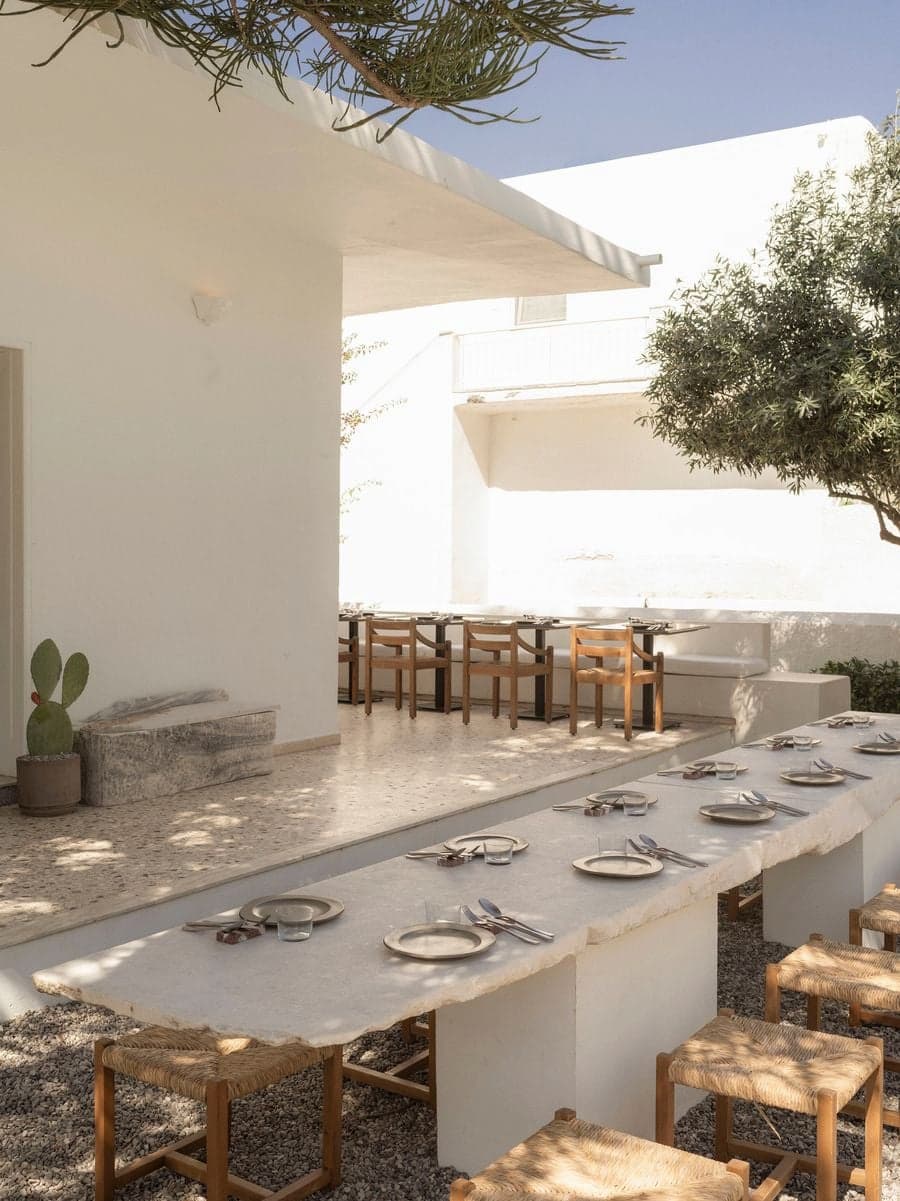 Communal marble dining table with woven oak stools in the sunlit Mediterranean courtyard of YAPA Antiparos, Greece