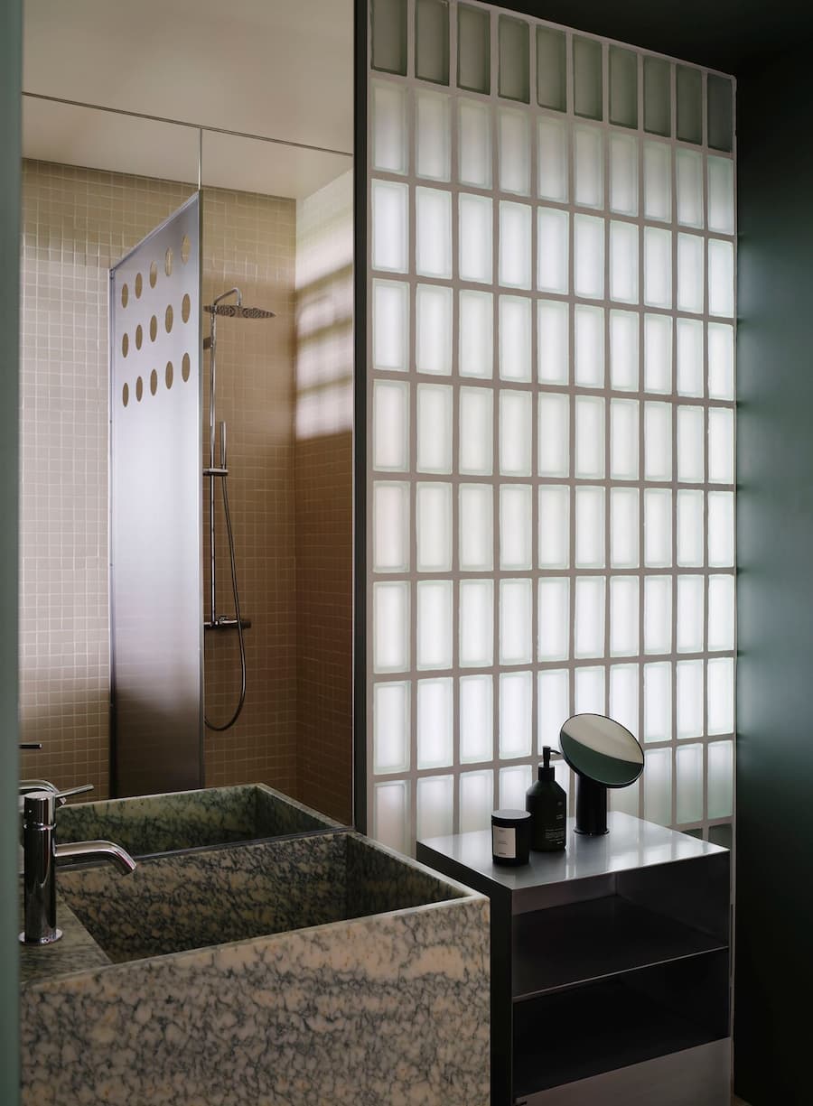 Bathroom combines glass block partition, marble sink and stainless steel shelving with soft natural light.