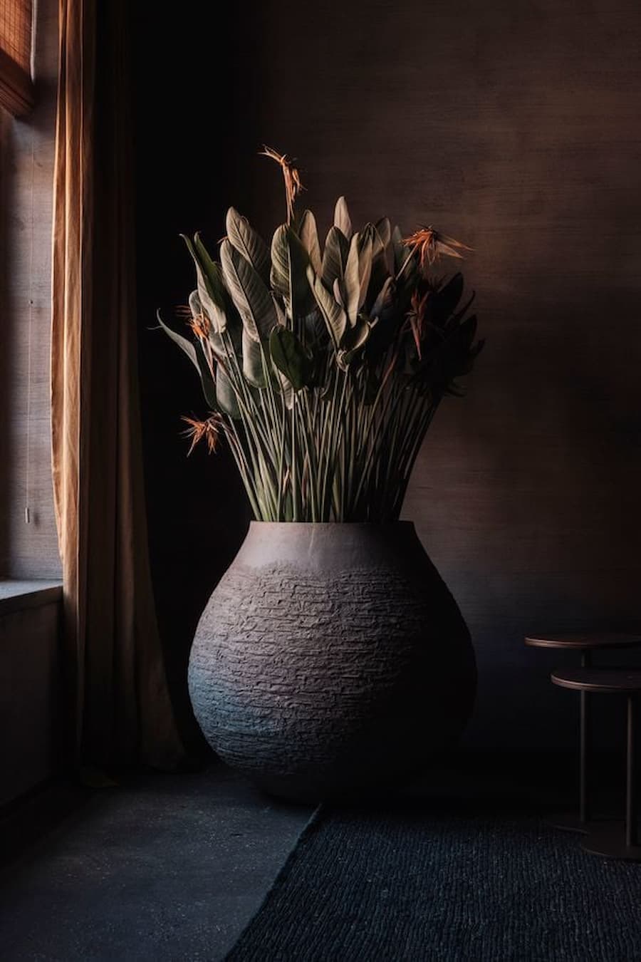 A large textured clay vessel filled with tropical foliage beside a dark interior wall
