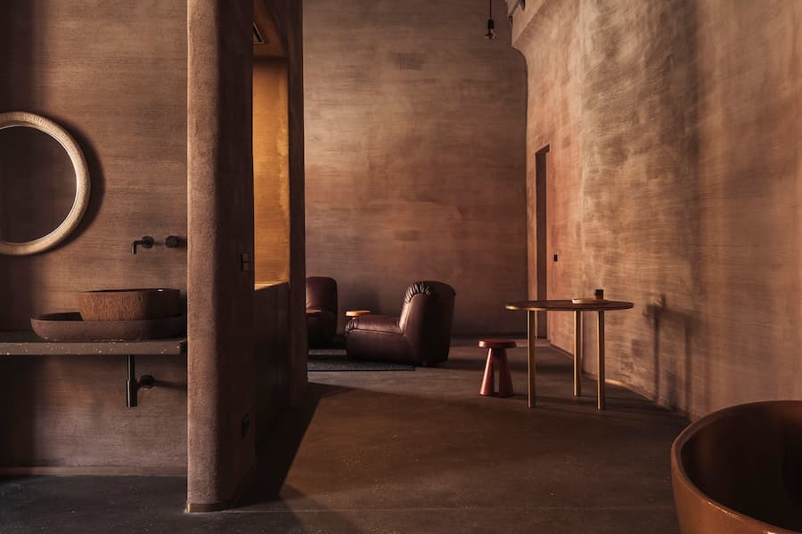 A clay washbasin and leather lounge chair share a warm interior shaped by textured earthen surfaces