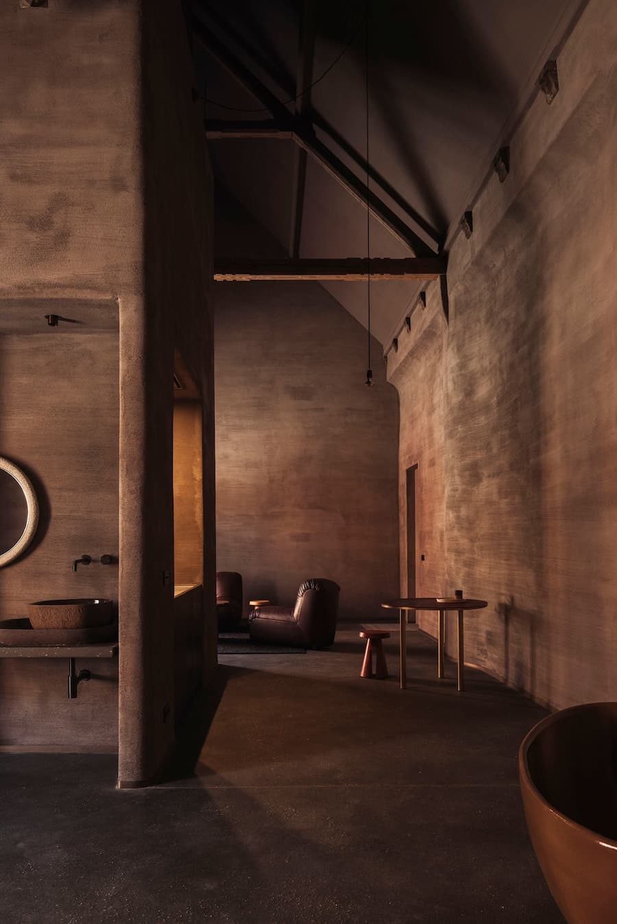 A tall earthen-plaster hall reveals exposed timber roof structure above a dark lounge and clay basin
