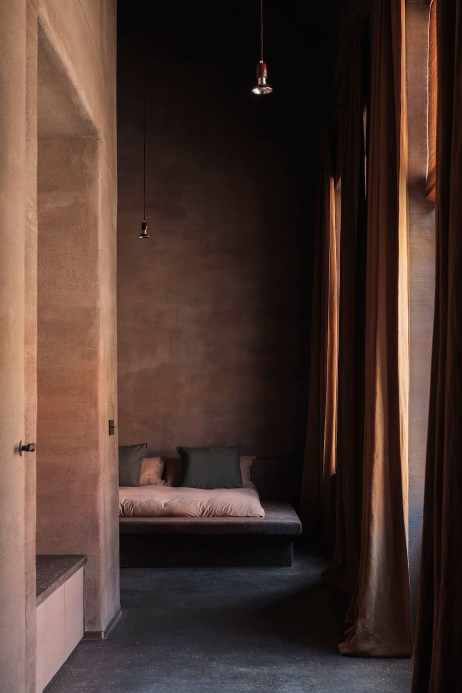A quiet bedroom sits behind heavy linen curtains with a low bed and suspended pendant lights