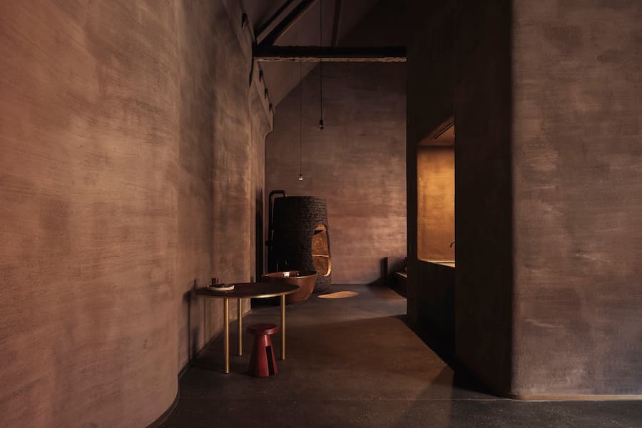 An elongated earth-toned corridor connects a round table and stool with a sculptural bathing area