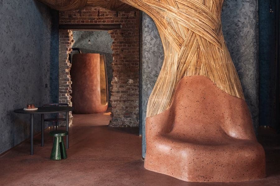 A sculptural terracotta lounge chair sits beneath an expressive woven canopy beside exposed brick