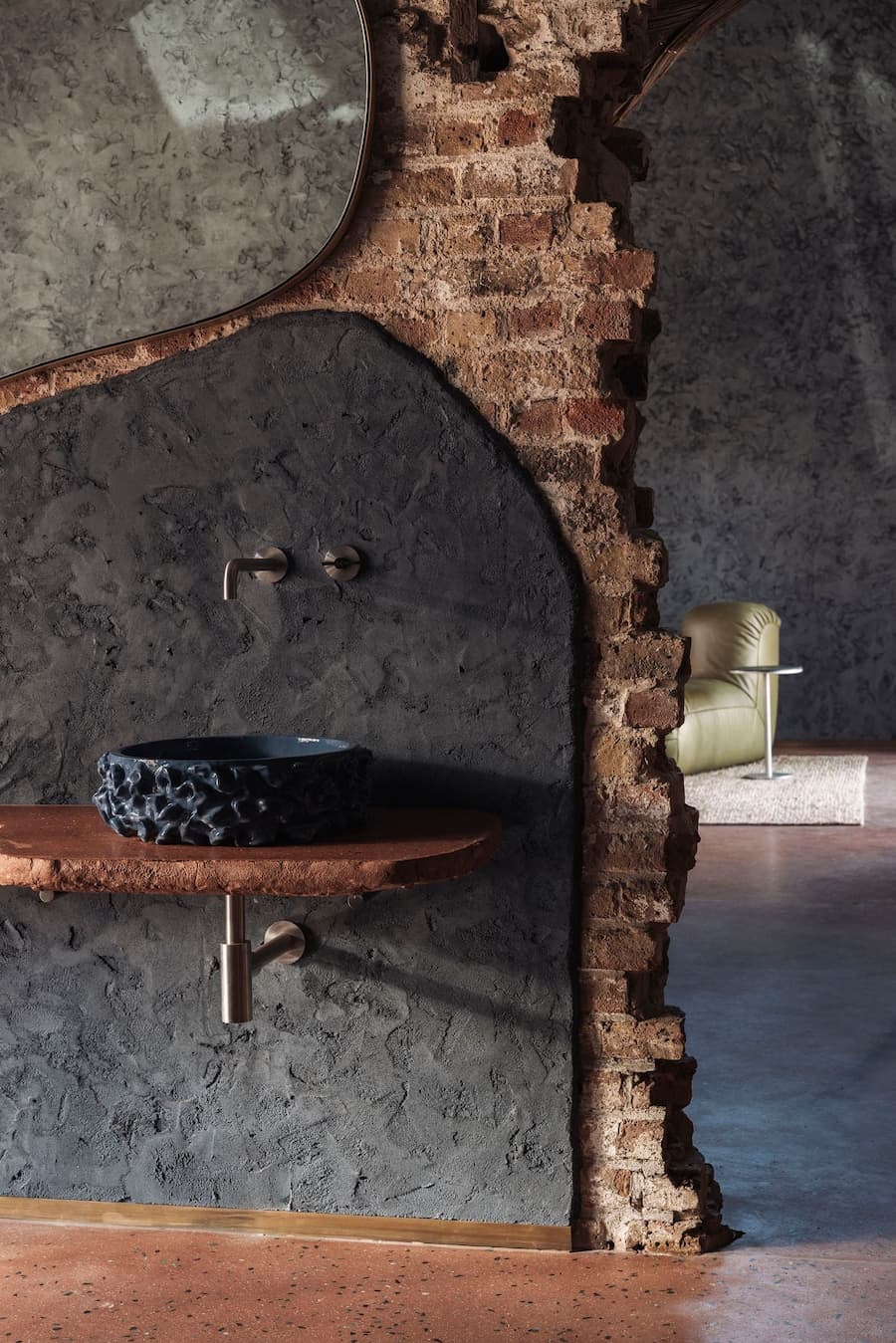 A dark ceramic washbasin rests on a rough timber shelf against a textured wall and exposed brick