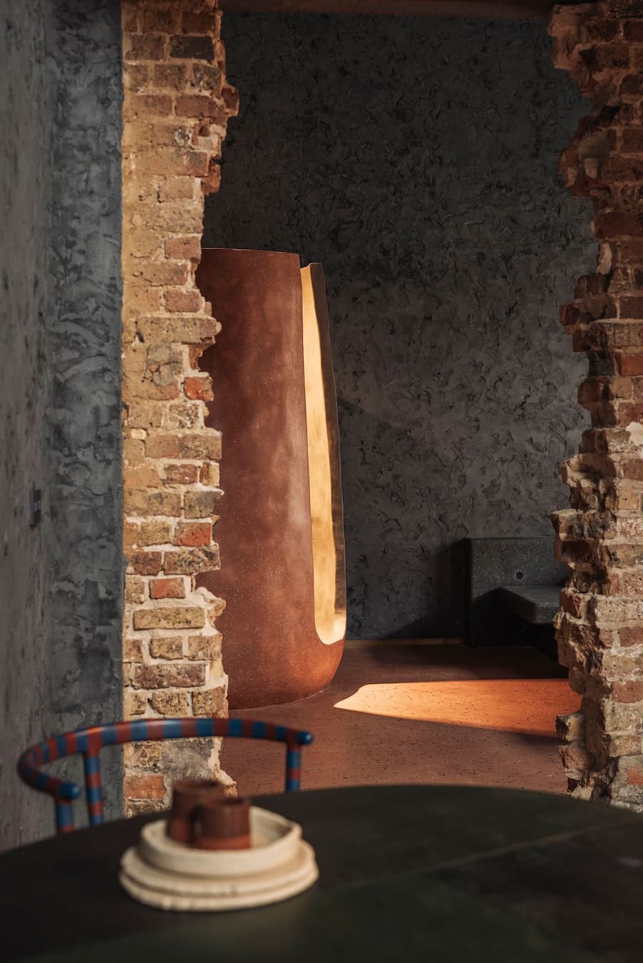 An exposed-brick opening frames a sculptural terracotta form against a dark textured plaster wall