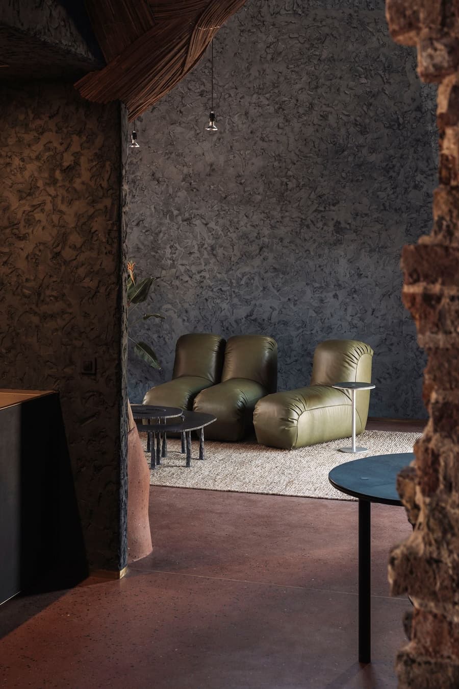 Olive leather lounge chairs sit beneath a woven ceiling detail beside rough plaster and exposed brick