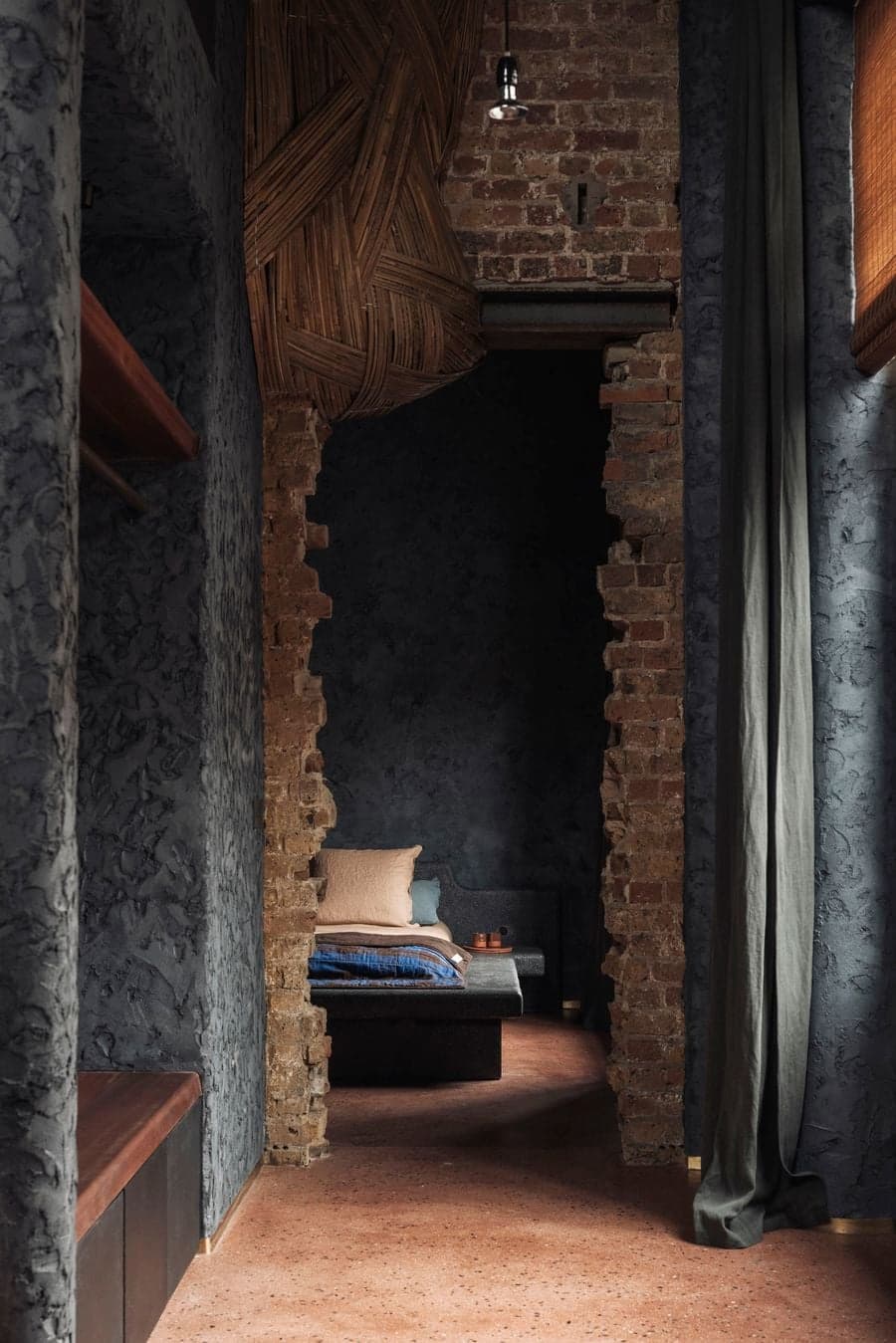 An exposed-brick threshold leads toward a dark bedroom framed by textured plaster and linen curtains