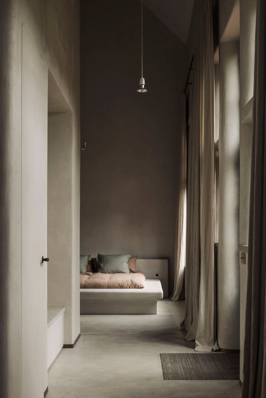 A quiet bedroom with soft plaster walls and tall linen curtains receives muted natural light