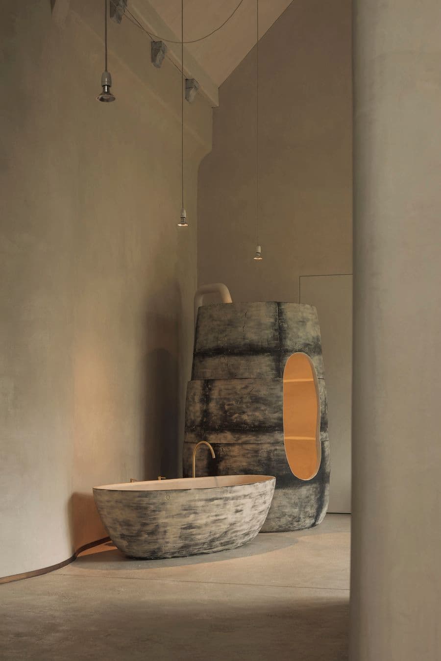 A weathered sculptural bathing structure stands behind a freestanding tub in a softly lit interior