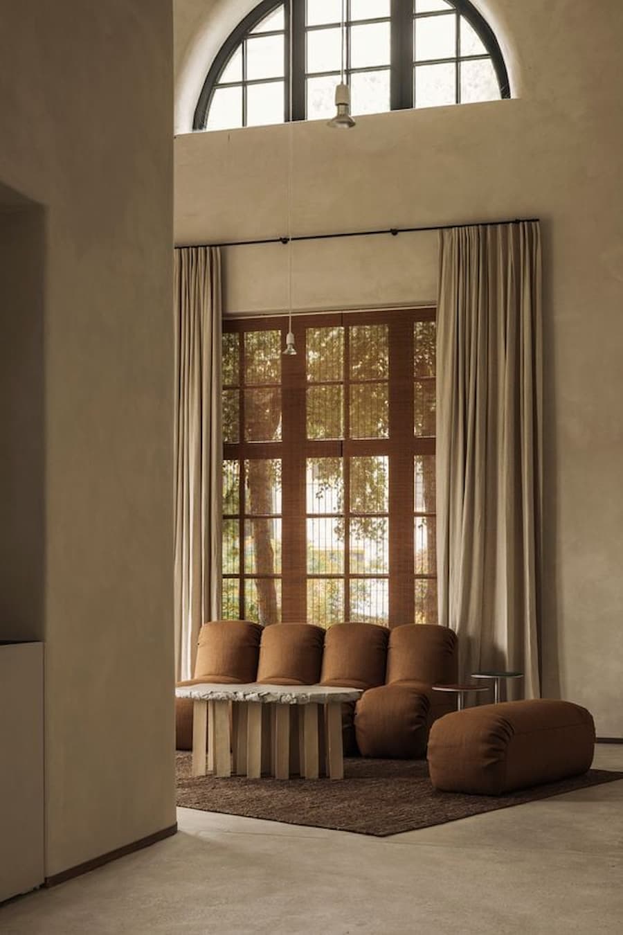 A large arched window overlooks a layered lounge with curtains, sculptural seating and soft daylight