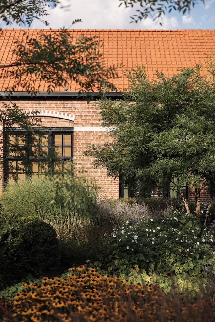 A brick house with a red tile roof emerges behind layered planting and ornamental grasses